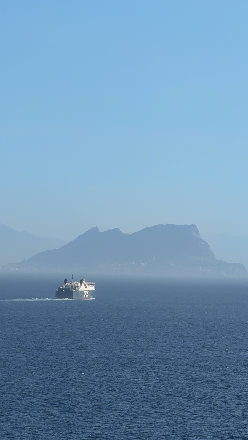 The Rock of Gibraltar seen across the Strait through heavy summer haze, its long ridge rising from a low spit of land and falling away in a sheer cliff at the right end, the whole silhouette flattened to pale blue-grey by the atmosphere. A band of white buildings is just visible along the shoreline beneath it. In the middle distance a black-hulled DFDS ro-ro ferry with a white superstructure crosses left to right, the DFDS wordmark large on its side, pushing a small white wake across otherwise calm deep blue water. Cloudless pale sky above.
