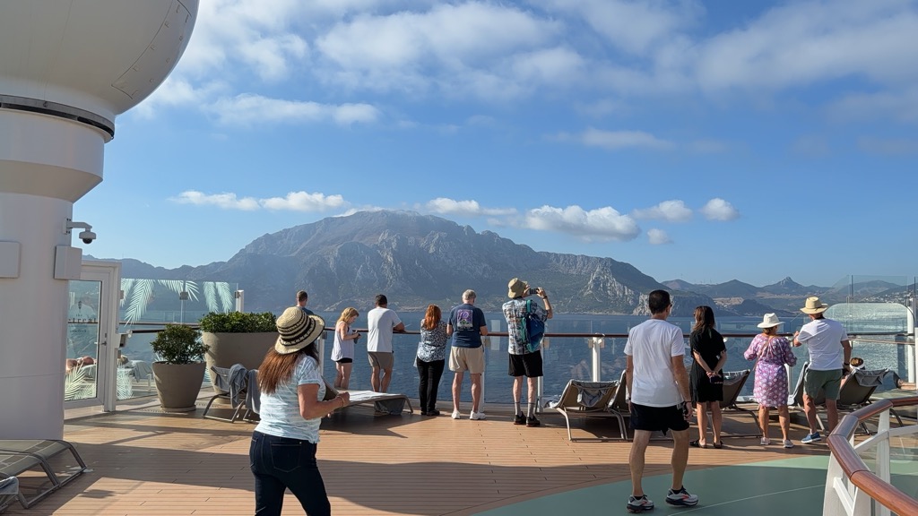 Passengers lined along the glass-and-steel railing of the ship's teak sun deck, looking out at the mountainous Moroccan coast across the Strait. The jagged ridge of Jebel Musa fills the horizon in grey-brown rock under scattered white cumulus and blue sky. About a dozen people watch from the rail -- several in straw sun hats, one in a bright floral shirt raising a phone to photograph, others in t-shirts and shorts -- while a woman in a white top and dark jeans walks across the foreground carrying a folded paper. Grey-cushioned loungers, potted palms in stone planters, and a white radar dome at the left frame the deck, which is paved in alternating teak-toned and pale green surfacing.