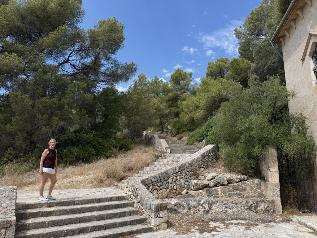 Sherri stands at the foot of a broad flight of rough stone steps climbing the pine-covered hillside toward the castle, turning back toward the camera. She wears a maroon sleeveless top, white shorts, and white sneakers. The steps rise from the cobbled terrace in the foreground and bend out of sight into the trees, bordered on the right by a dry-stone retaining wall with the corner of the chapel and its iron gate at the frame edge. Dry yellow grass, low scrub, and Aleppo pines fill the slope under a bright blue sky with a few thin white clouds.