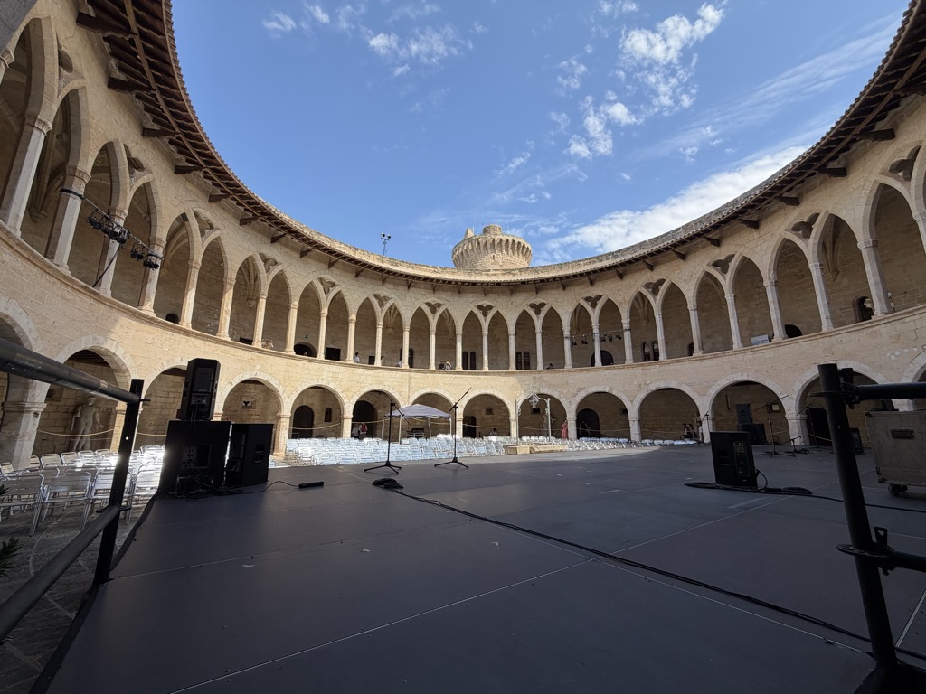 The circular courtyard at the heart of Castell de Bellver, shot from the edge of a temporary black stage floor. Two tiers of arcades ring the whole space -- rounded arches on square piers at ground level, taller pointed Gothic arches on slender double columns above -- all in pale honey limestone, with the castle's freestanding keep visible over the roofline at the far side. Rows of white folding chairs are set out on the far half of the yard, and stage gear fills the foreground: black monitor wedges, microphone stands, cabling taped down, lighting bars rigged under the upper arcade at the left, and a white canopy against the far wall. Deep blue sky with wisps of cloud above.