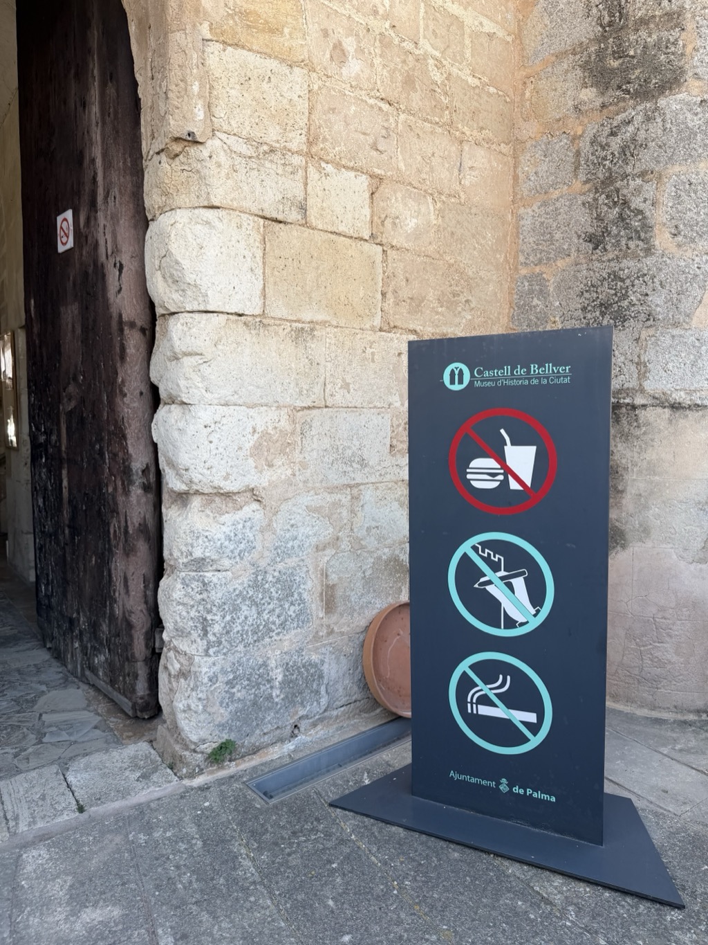 A dark grey A-frame sign standing on the worn stone threshold of the castle's deep entrance archway, beside a heavy studded timber door. The panel is headed Castell de Bellver, Museu d'Historia de la Ciutat in white and pale green, below which three pictograms are struck through: a burger and soft drink cup in a red prohibition circle, a figure climbing the battlements, and a lit cigarette. The footer reads Ajuntament de Palma with the city crest. The surrounding walls are pale ashlar block, weathered and patched, with a terracotta saucer resting on the ground behind the sign.