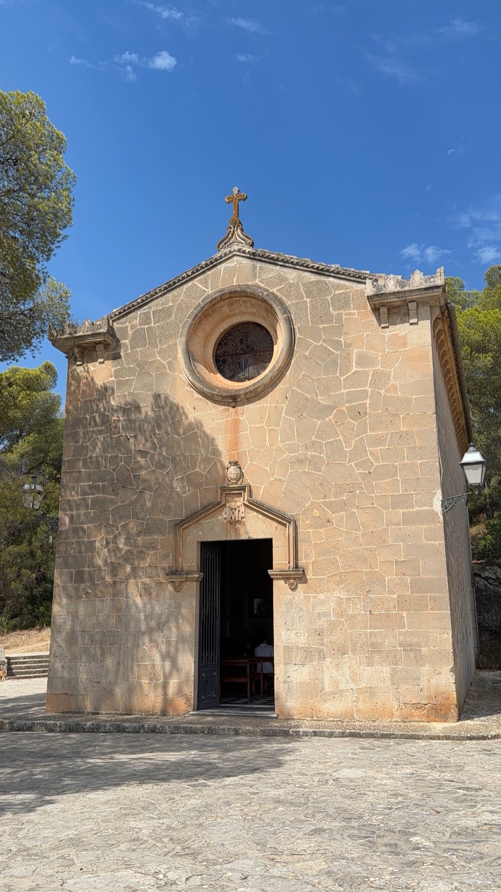 The front of a small stone chapel on the wooded hillside below the castle, shot straight on from the cobbled forecourt. The honey-colored limestone facade rises to a gable topped with an ornate wrought-iron cross, with a round rose window of tracery glass at its center and a pointed stone doorway below carrying a carved Marian monogram. The heavy iron gate stands open, and inside a person in white can be seen sitting in the wooden pews. Wrought-iron lanterns are bracketed to the walls at either side, pines press in from both edges of the frame, and the sky above is cloudless deep blue.