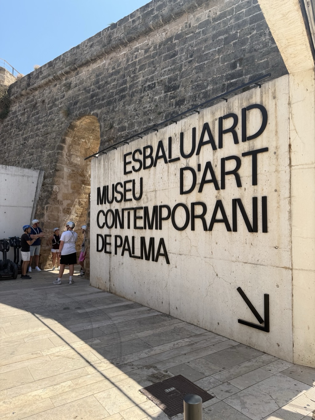 The entrance wall of Es Baluard, shot at an angle from the paved forecourt. A tall white concrete slab carries the museum's name in large black cut-out letters -- ESBALUARD MUSEU D'ART CONTEMPORANI DE PALMA -- with a black arrow at the lower right pointing down toward the entrance. Behind and above it rises the massive dark stone rampart of the old city bastion, with a rounded arch tunnel cut through it at the left. In the shade beside the tunnel, several people from the Segway tour stand talking in white safety helmets, their Segways parked along the wall. Bright sun rakes across the pale stone paving in the foreground, with a manhole cover and a steel bollard at the bottom of the frame.