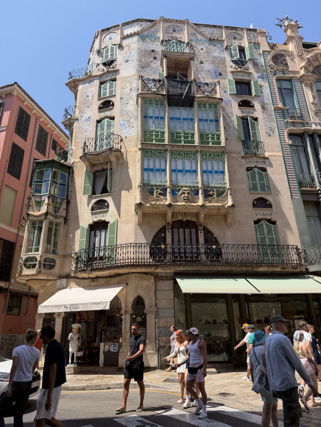 A five-story Palma modernista building on a street corner, shot from below against a cloudless blue sky. The upper facade is encrusted with pale mosaic and floral relief work; green-shuttered windows and slender wrought-iron balconies wrap the rounded corner, and a broad glazed gallery of green-framed windows fills the center, lettered CLINICA DENTAL across the glass. A carved grotesque face with flanking sea creatures sits above the second-floor balcony, which runs the width of the building on ornate ironwork. At street level, a cream awning marked St. Cristo shades a shopfront where a life-size chef figure stands beside the door, with a jewelry shop under a pale green awning to the right. A crowd of summer pedestrians in shorts, sundresses, and caps crosses the zebra-striped street in the foreground; a pink-washed building adjoins at the left.