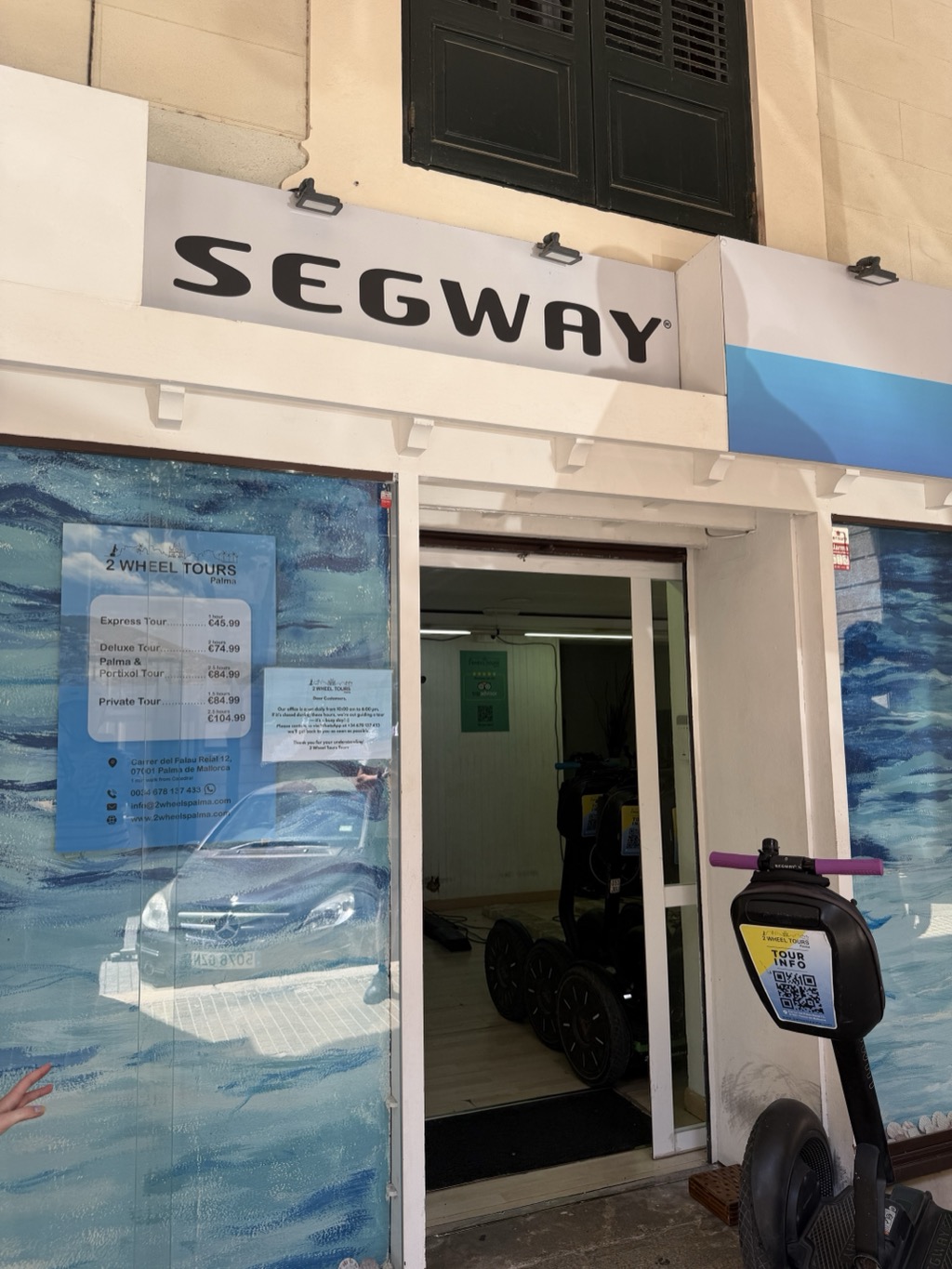 The storefront of 2 Wheel Tours Palma on a narrow old-town street, with a large white SEGWAY sign mounted above the entrance. The glass frontage is printed with a wash of blue and white water, and a price list is taped inside reading Express Tour 1 hour 45,99 EUR, Deluxe Tour 2 hours 74,99 EUR, Palma and Portixol Tour 84,99 EUR, Private Tour 84,99 to 104,99 EUR, with the address Carrer del Palau Reial 12, 07001 Palma de Mallorca and contact details. Through the open door, several black Segways are parked in a bare tiled room. A Segway stands at the right of the frame in the foreground, a yellow TOUR INFO placard with a QR code clipped to its handlebar column. Dark green shutters and pale render fill the upper facade.