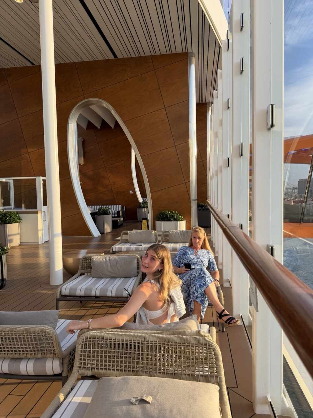 Two women on the ship's shaded outer deck at golden hour, seated on rattan loungers with striped cushions. In the foreground a young blonde woman in a white top turns back toward the camera with one arm along the seat back; behind her an older woman in a blue and white paisley dress sits holding a phone. The deck is panelled in warm timber with a tall white sculptural arch framing a lounge area behind, potted plants in white planters along the wall, and a polished wooden handrail running the glass balustrade at the right, beyond which the orange hull of the Magic Carpet and the port cranes are visible.