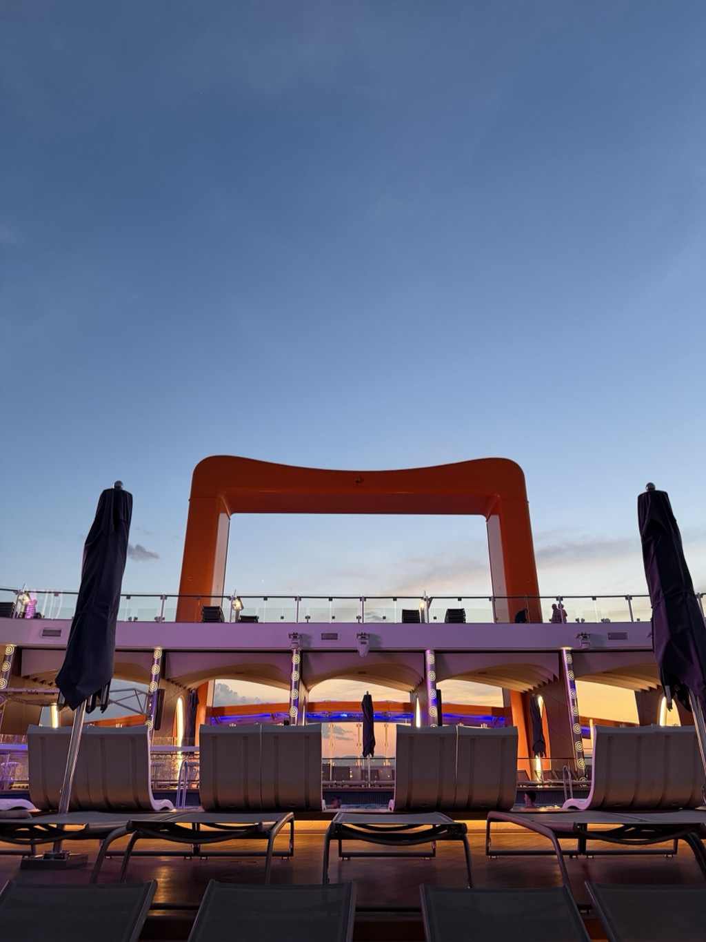 The ship's pool deck at dusk, shot from between empty loungers. The bright orange frame of the Magic Carpet platform rises against a deep blue twilight sky streaked with pale cloud, silhouetted above the upper deck's glass rail. Below it the sheltered pool area glows with purple and amber light under scalloped white canopies, a strip of blue-lit water visible through the openings. Furled navy parasols stand at either side of the frame, and rows of grey sun loungers fill the foreground on the darkened teak deck.