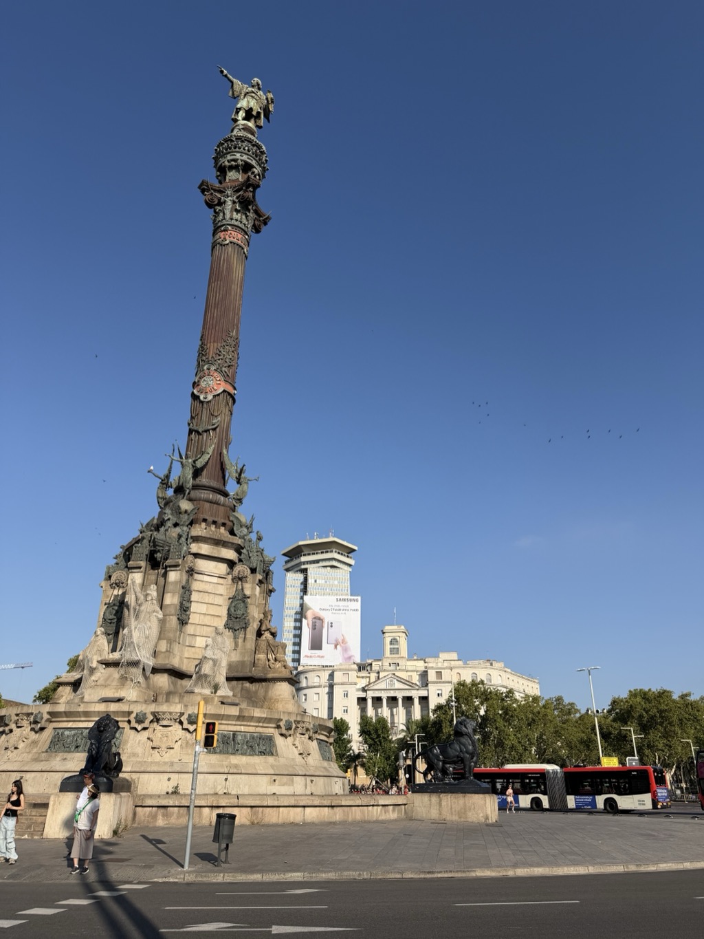 The Columbus Monument at the foot of La Rambla, shot from across the road against a deep blue sky. The bronze figure of Columbus stands atop a tall fluted column pointing out to sea, the base ringed with bronze allegorical figures and stone lions on plinths, with protective mesh over part of the stonework. Behind it are a glass-fronted tower block carrying a large Samsung Galaxy billboard and the classical stone facade of the port authority building. Buses and a few pedestrians cross the wide junction in the foreground, and birds scatter across the empty sky at the right.