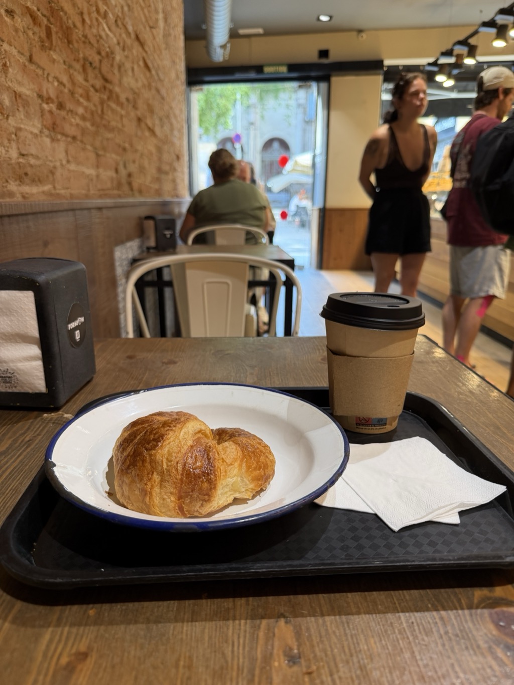 A breakfast tray on a wooden cafe table: a plain croissant on a white enamel plate with a blue rim, a takeaway coffee cup with a black lid and kraft sleeve, and a folded paper napkin. A black napkin dispenser sits at the left against an exposed brick wall. Behind, the cafe's glass frontage looks out to a sunlit street where a red-and-white bollard stands; a person in an olive t-shirt sits at a table by the window, and two customers wait at the counter at the right under track lighting.