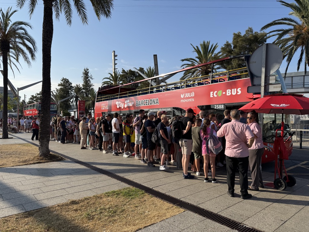 A long queue of cruise passengers waiting along a palm-lined pavement beside a red City Tour Barcelona double-decker bus, number 2588, its side lettered City Tour BARCELONA and barcelona.city-tour.com with a JULIA moventis logo and a green ECO-BUS badge. The open top deck shows rows of empty red seats. People in shorts, t-shirts, sun hats and backpacks stand in line under tall palms against a cloudless blue sky, with a second red bus waiting further back. At the right two staff in pink shirts stand beside a red City Tour umbrella and a loaded hand truck.