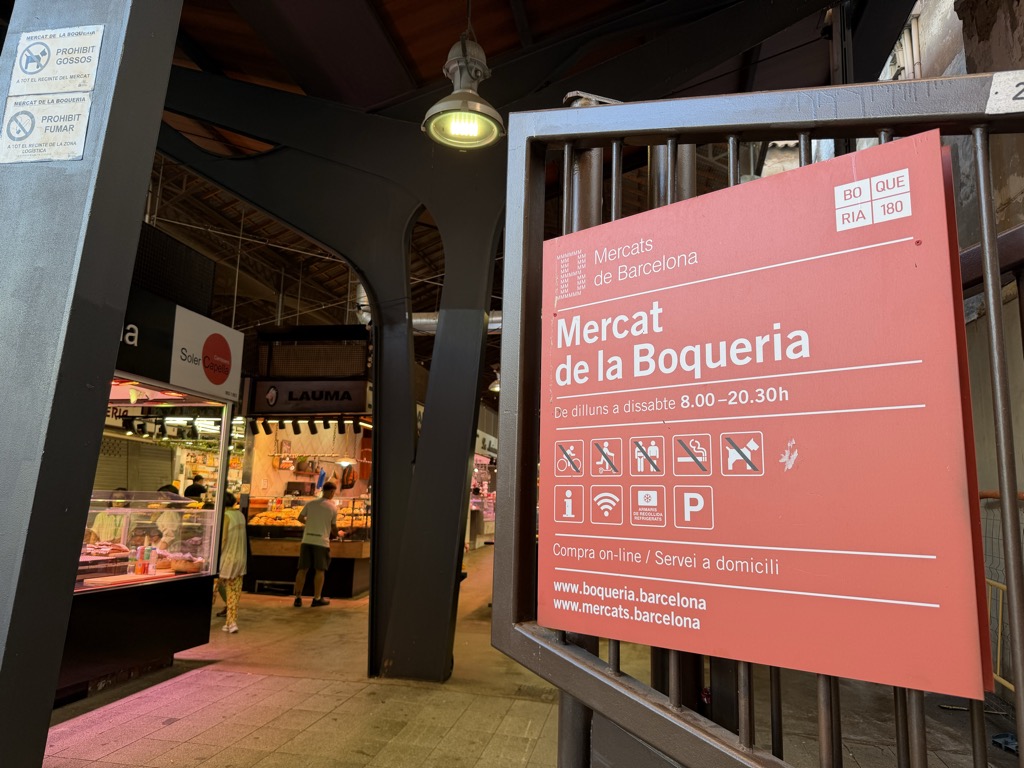 A large coral-pink sign in a metal frame at a side entrance to the market, headed Mercats de Barcelona and Mercat de la Boqueria in white, with opening hours De dilluns a dissabte 8.00-20.30h. Two rows of pictograms below prohibit bicycles, scooters, smoking and dogs alongside information, wi-fi and parking symbols, and the footer gives www.boqueria.barcelona and www.mercats.barcelona. A small panel at the top right reads BOQUERIA 180. Through the dark steel-columned entry behind it, a lit butcher's counter and a few shoppers are visible under the market's timber roof.