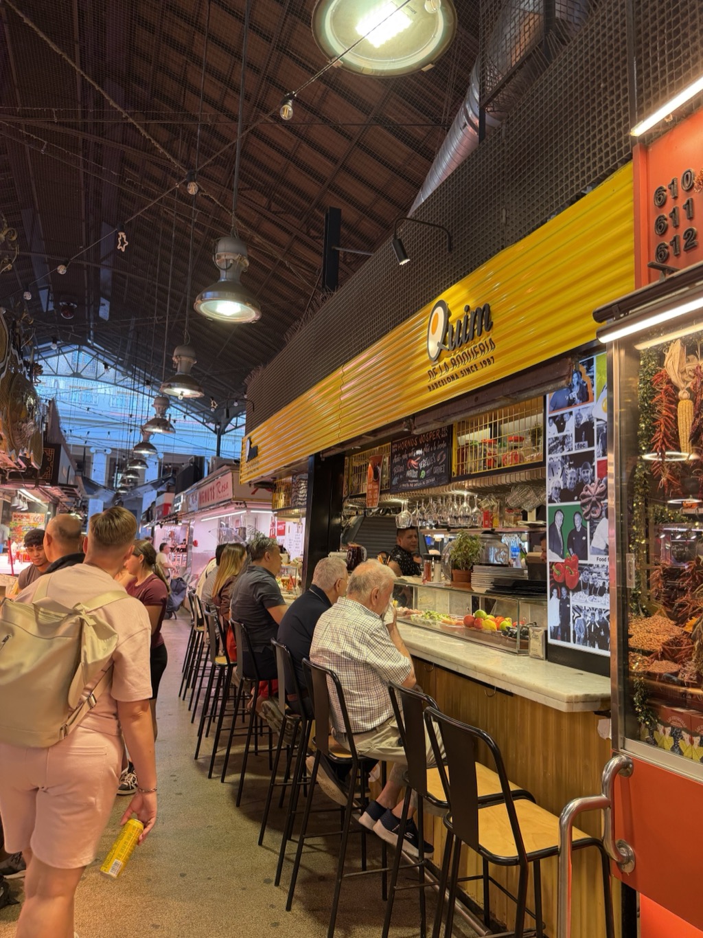 Inside Mercat de la Boqueria, looking down an aisle toward the Quim de la Boqueria stall, its curved yellow signboard lettered Quim de la Boqueria, Barcelona since 1987, with stall numbers 610 611 612 on an orange panel at the right. Diners sit on black stools along the marble counter, an older man in a checked shirt and shorts nearest the camera, while a cook works behind the bar under hanging glassware and a chalkboard menu. Strings of dried red peppers hang in the glass case at the right. Industrial pendant lamps drop from the high timber and steel roof, and shoppers move down the aisle at the left.