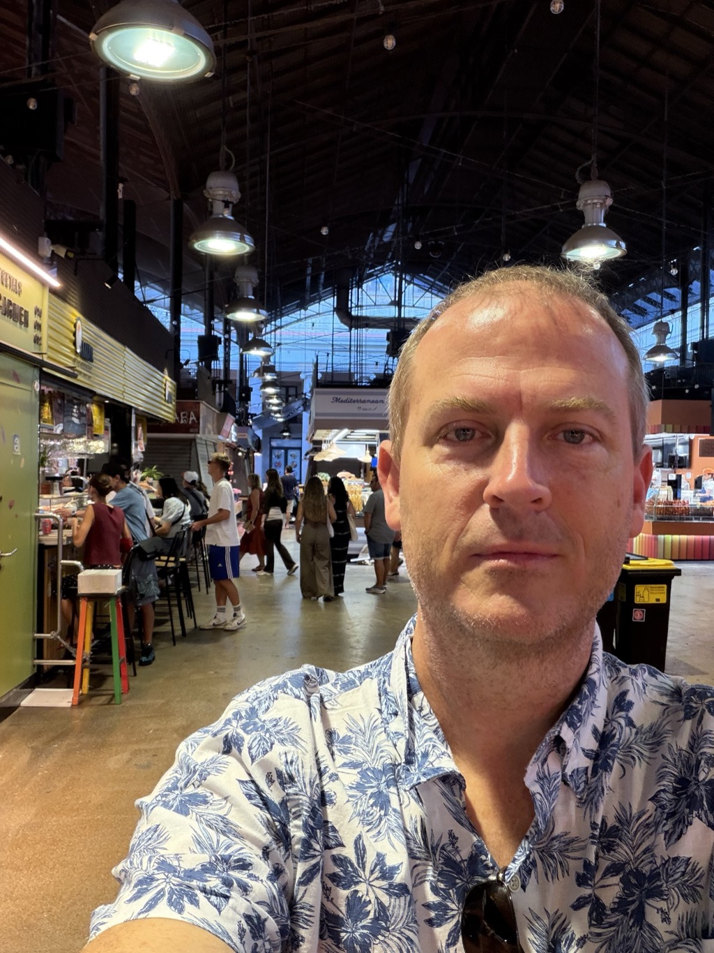 Brian takes a selfie in the aisle of Mercat de la Boqueria, filling the right of the frame. He wears a white short-sleeved shirt printed with large blue-grey leaves and sunglasses hooked on the placket, and looks tired and flushed, not smiling. Behind him the market hall recedes under its arched roof -- pendant lamps on long cords, a stall sign reading Mediterranean, shoppers crossing the polished concrete floor, and diners on brightly coloured stools at a counter to the left. A yellow waste bin stands against the stall at the right.