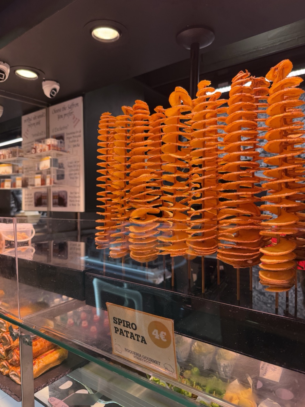A market stall display at the Boqueria holding tall skewers of spiral-cut potato crisps, deep orange and stacked in ruffled columns behind glass. A card propped in front reads SPIRO PATATA 4 euro, BOQUERIA GOURMET STREET FOOD. Spotlights and a small security camera are mounted on the dark ceiling above. A lower case at the front holds cured meats, stuffed peppers and green olives, and a shelf of packaged saffron with handwritten signs stands behind the glass at the left.