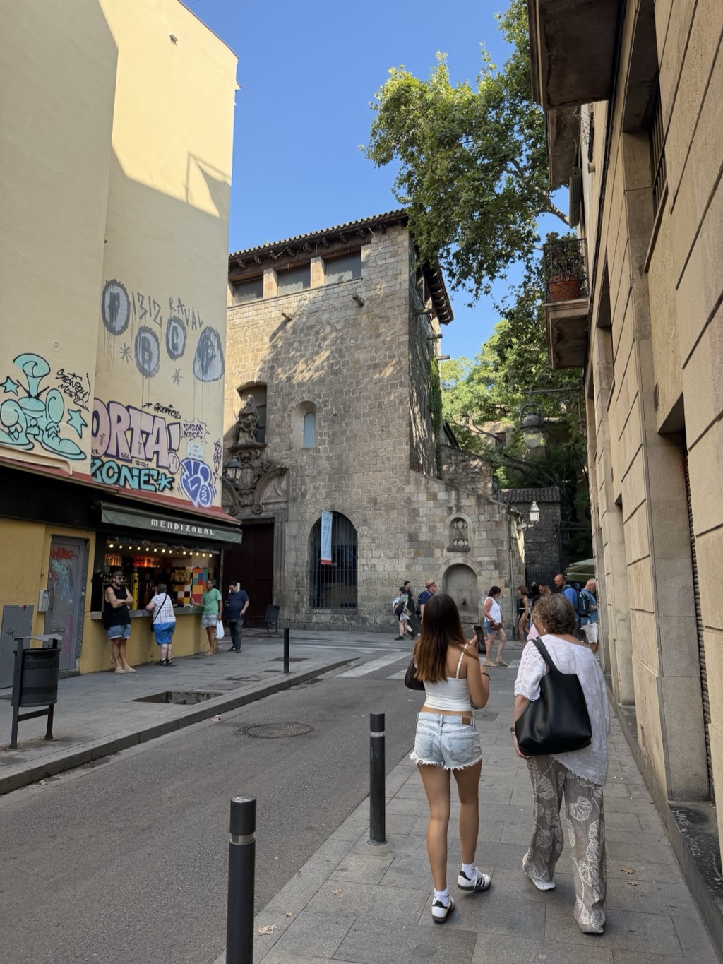 A narrow Raval street corner in bright morning sun. At the left a small kiosk bar under a green awning lettered MENDIZABAL, its front tiled in coloured squares and hung with festoon lights, a few people standing at the counter; the pale wall above is covered in graffiti tags and a large mural. Ahead stands a plain medieval stone building with a small arched window and an iron-grilled doorway, a tree spilling over its roofline and an ornate baroque stone portal at its left. Two women walk away from the camera along the pavement at the right, one in a white top and denim shorts holding a phone, the other in a white blouse and patterned trousers with a black tote bag.