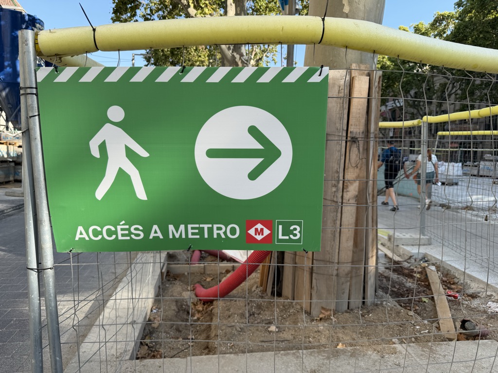 A green temporary pedestrian sign zip-tied to construction fencing on La Rambla, reading ACCES A METRO with a walking figure, a white arrow in a circle, and the red Barcelona metro M logo beside L3. Yellow flexible ducting runs above the sign along the hoarding. Behind the wire mesh, a plane tree trunk is boxed in with timber boards, red conduit snakes through excavated bare earth, and stacked materials and white bags sit along the dug-up promenade. A couple of pedestrians pass on the narrowed walkway beyond.