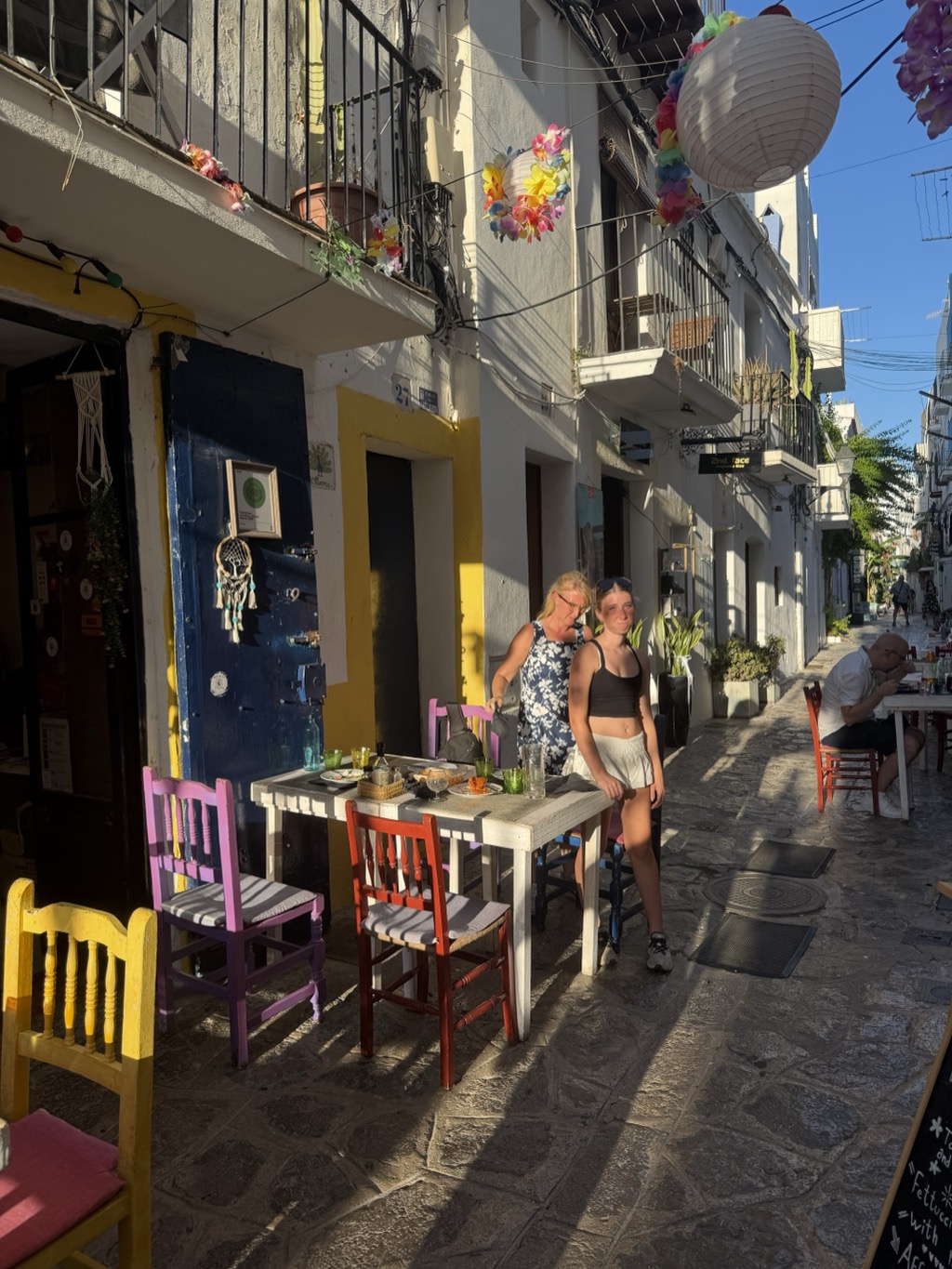 A narrow cobbled alley in the old town in low evening sun, lined with pavement tables and brightly painted wooden chairs in yellow, pink, purple, red and blue. At a white table in the foreground a woman in a navy and white floral dress leans over the table setting something down, while a young woman in a black crop top, white skirt and sunglasses on her head stands beside it looking toward the camera; the table holds glasses, a small dish and a bottle. A deep blue studded door with a dreamcatcher hanging beside it fills the left of the frame, and paper lanterns and bright fabric flower garlands are strung overhead between the balconies. Further down the alley a man in a white shirt sits reading at another table, with whitewashed walls and a chalkboard menu beyond.