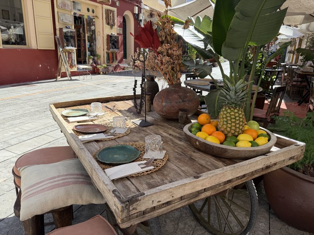 A restaurant table built from an old wooden cart with a spoked wheel, set for three on a sunlit pedestrian street. Woven rattan placemats hold glazed ceramic plates in green and terracotta, cut-glass tumblers and rolled napkins with cutlery. At the far end a shallow wooden bowl is piled with oranges, lemons, limes and a whole pineapple, beside a round terracotta urn holding dried flowers and a tall wrought-iron candle stand. A banana plant and potted greenery crowd in at the right under a white parasol, and cushioned stools with a striped linen cushion sit at the left. Across the street a red-washed building houses a small shop hung with jewellery and macrame.