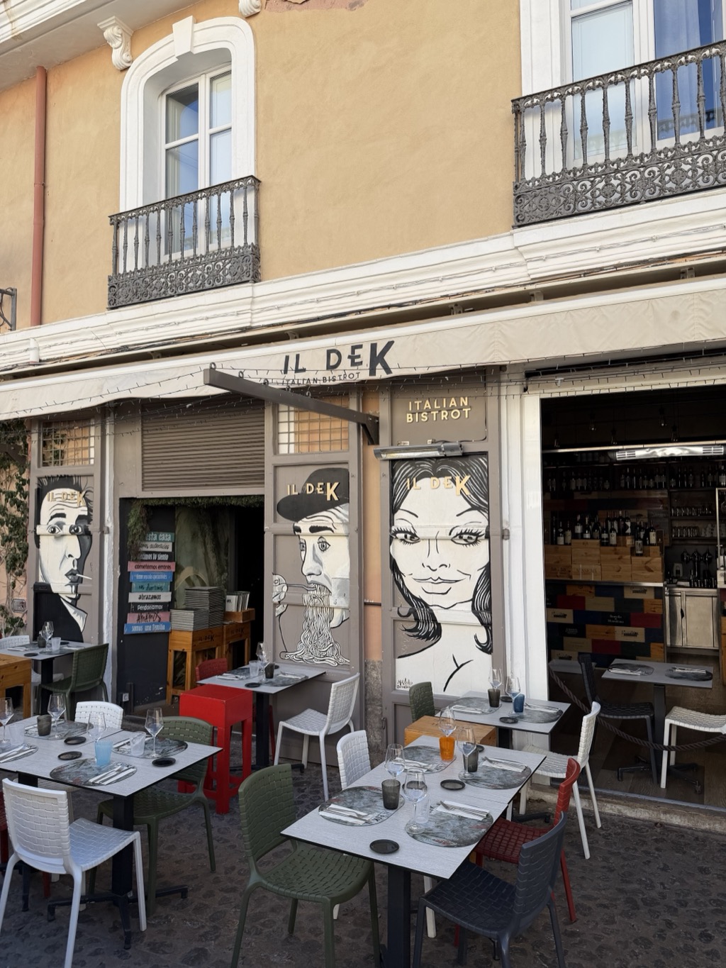 The frontage of Il DeK Italian Bistrot on an old-town street, its cream awning lettered IL DeK with ITALIAN BISTROT beneath and repeated in gold on the wall. Three large black-and-white mural portraits are painted on the shutters and panels -- a man in a hat smoking, a man in a flat cap slurping a strand of spaghetti, and a woman with long dark hair and heavy eyeliner. Through the open doorway at the right, a bar is stacked with wine bottles and wooden crates. Pavement tables in grey are laid with wine glasses, cutlery and coloured tumblers, surrounded by mismatched chairs in white, olive green, red and grey, with a bright red high table at the centre. Above, an ochre facade carries two tall windows with ornate wrought-iron balconies.