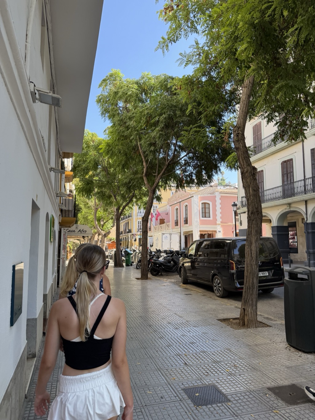 A young woman in a black crop top, white skirt and sunglasses pushed into her ponytail walks away from the camera down a shaded old-town street. The whitewashed wall of a building runs along the left with a small shopfront awning lettered Casa Afro further down. Leafy trees arch over the pavement, and beyond them a row of pastel buildings in cream, yellow and dusty pink stand in bright sun, one with a small flag hanging from a balcony. Parked scooters line the far kerb beside a black Mercedes van, and a green wheelie bin and dark litter bin stand on the tiled pavement. Deep blue cloudless sky above.