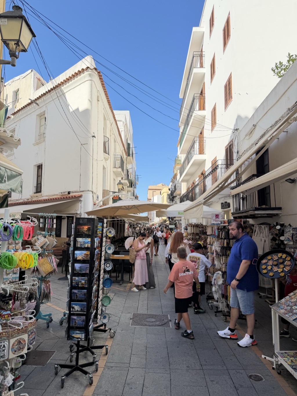 A narrow, busy shopping street in the old town, walled by tall white buildings with green and brown louvred shutters and iron balconies. Souvenir displays crowd both sides -- a rotating rack of postcards, hoops of coloured bangles, keyrings, printed ceramic plates, straw bags and racks of jewellery, with a tray of fridge magnets held out on a stand at the right. A man in a blue polo shirt and denim shorts browses at the right; two boys in a salmon t-shirt and a white t-shirt walk down the middle, and a woman in a pink dress checks her phone beside a woman in cream. Cream parasols shade cafe tables further down, and a wrought-iron street lantern hangs at the top left against deep blue sky.