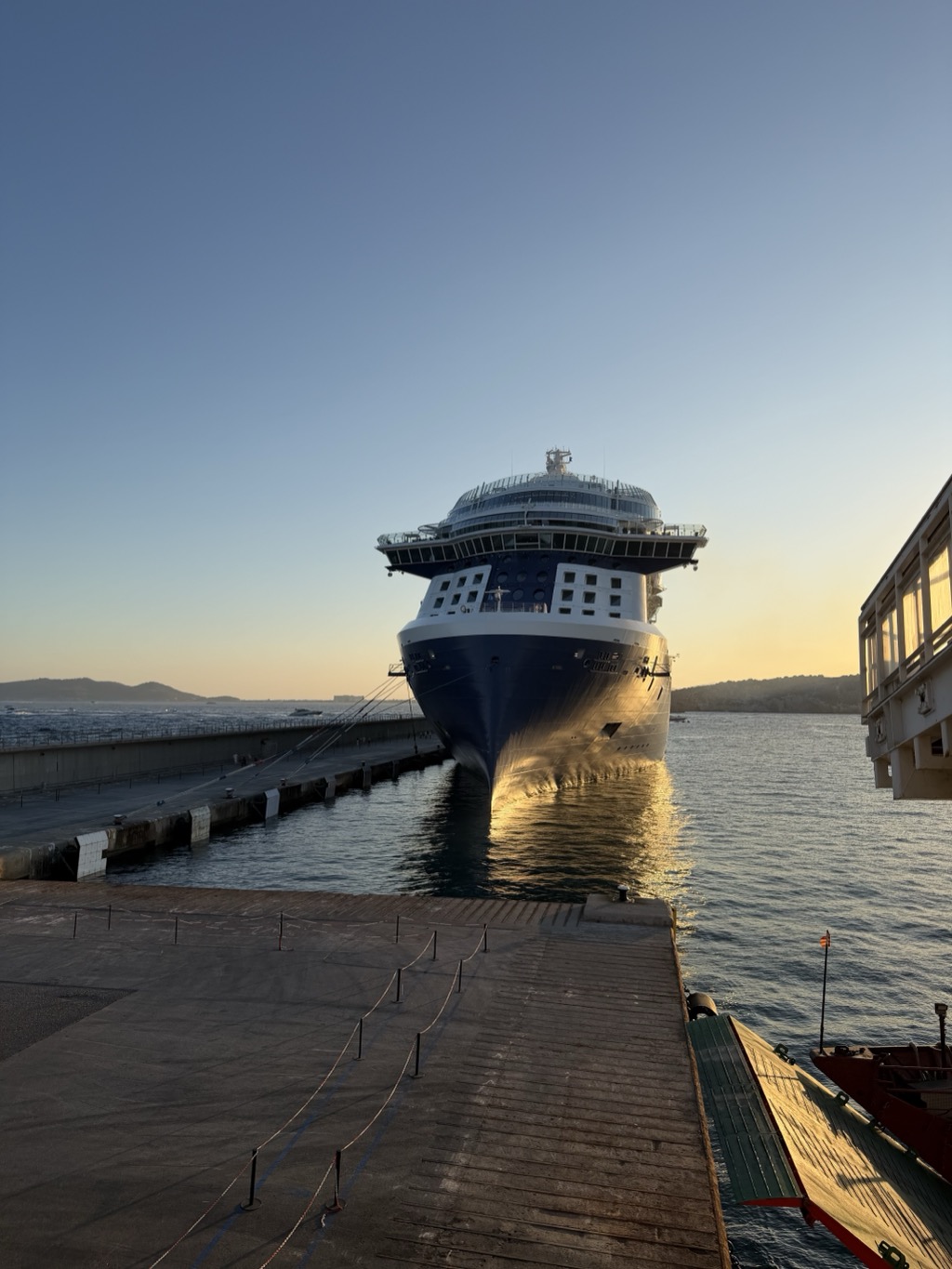 The Celebrity Apex viewed head-on from the pier at sunset, its dark blue bow rising above its own reflection in still water, mooring lines running down to bollards along the concrete quay at the left. The white superstructure and the glass-walled observation lounge catch the last warm light. A stanchion-and-rope barrier zigzags across the wooden decking in the foreground, a small red tender and a green gangway structure sit at the right, and low hills and moored boats are visible on both sides of the harbour under a clear sky graduating from blue to gold.