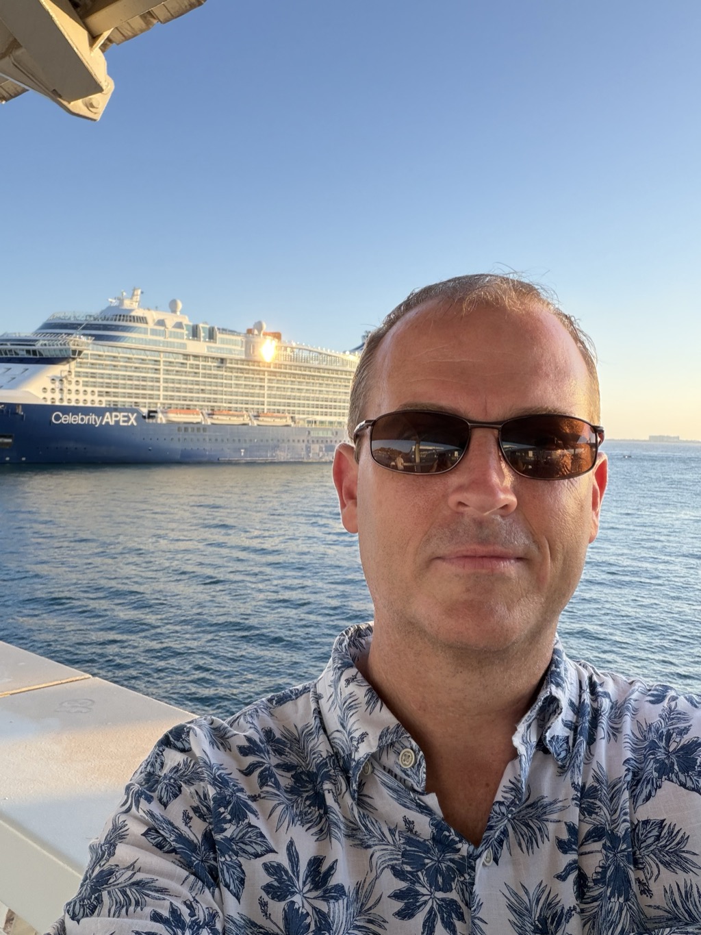 Brian takes a selfie on the quayside at golden hour, filling the right of the frame. He wears dark wraparound sunglasses and a white short-sleeved shirt printed with large blue palm leaves, and looks into the camera with a slight smile. Behind him across calm blue water the Celebrity Apex lies moored, its navy hull lettered Celebrity APEX and the low sun flaring off a window on the upper deck. A pale cream awning edge juts into the top left corner and a white rail runs along the bottom left. The sky fades from blue to warm gold at the horizon.