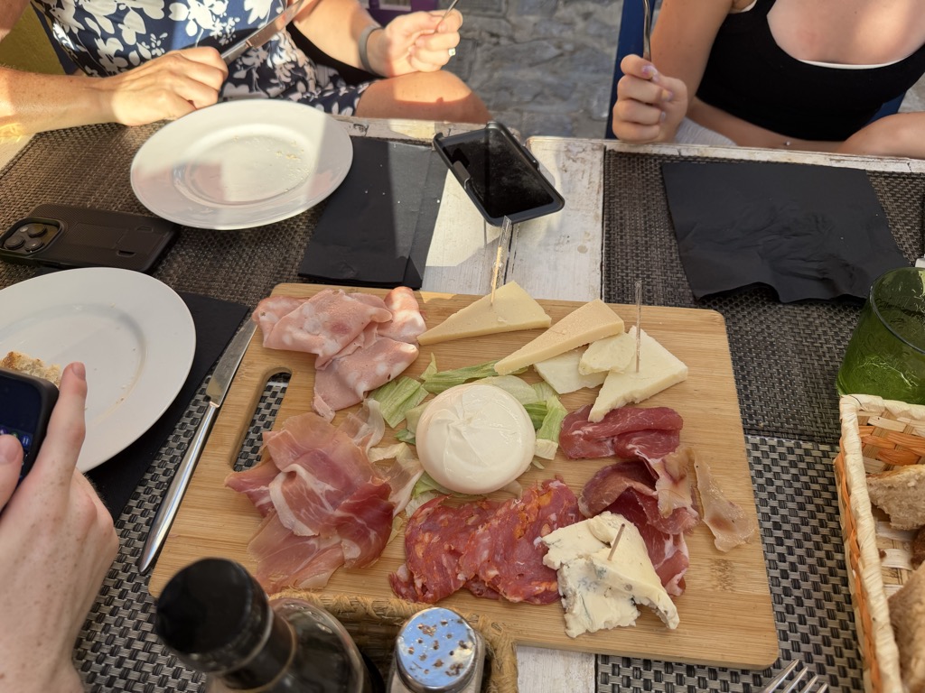 A wooden charcuterie board on a restaurant table, laid with folded slices of prosciutto, coppa and a dark chilli-flecked salami, wedges of hard yellow cheese, a crumbling white cheese and a blue-veined cheese pinned with cocktail sticks, arranged around a whole round ball of creamy burrata on a bed of shredded lettuce. A basket of bread sits at the right beside a green tumbler, with white side plates, black napkins, a bottle and a shaker around the edge of the frame. Three pairs of hands and forearms are visible at the far side of the table -- one in a navy and white floral dress, one bare, one in a black top -- with phones resting on the placemats.