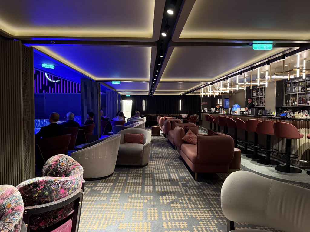 A wider view down the same lounge toward its bar. Recessed ceiling panels are lit warm gold with deep blue cove lighting between them, and green illuminated exit signs hang from the beams. On the right, a long bar runs the length of the wall with backlit vertical light fixtures, tiered shelves of liquor bottles, and a row of rust-red barstools with black bases; two bartenders in dark uniforms work behind it. Rust-red banquettes and grey tub chairs line the floor, one foreground chair upholstered in a bright multicolored floral print. At the far left a handful of guests sit in a blue-lit alcove beside a neon wall sign. The carpet is grey with a gold houndstooth pattern.