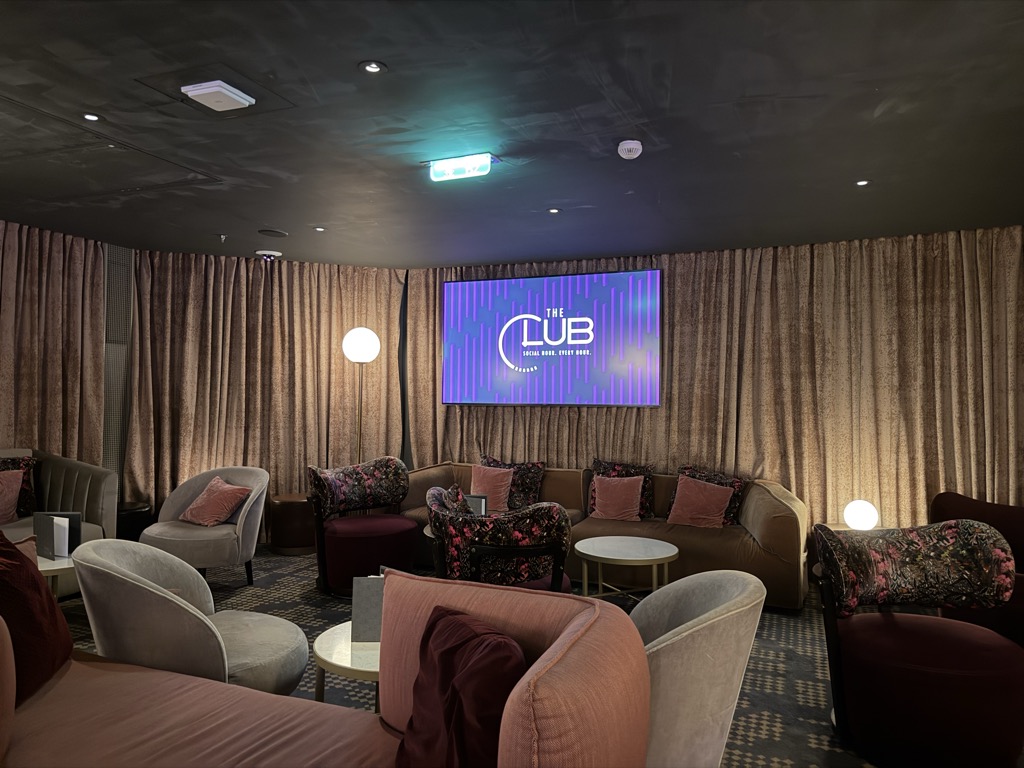 A dim lounge aboard the ship with a dark grey ceiling, floor-to-ceiling gold-beige curtains, and a large wall-mounted screen showing a purple and green graphic reading THE CLUB — SOCIAL HOUR, EVERY HOUR. Grouped seating fills the room: pale grey and blush tub chairs, a tan curved banquette with mauve and floral cushions, burgundy velvet armchairs, and round white marble-topped cocktail tables. Two globe floor lamps glow, and a small teal stage light is fixed to the ceiling. The carpet is dark blue with a pale geometric pattern. The room is empty of people.