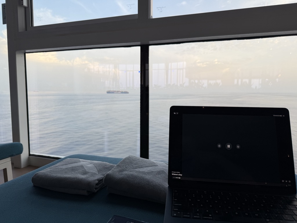 A view from a blue upholstered window seat aboard the ship, looking out through a tall glass wall at a flat pale sea under a hazy sky streaked with thin cloud. A container ship sits small on the horizon. Two folded grey towels rest on the bench, and an iPad in a keyboard case stands open on the right with a dark audiobook player screen showing pause and skip controls and the title Widow's Bay. Reflections of the interior are visible in the glass.