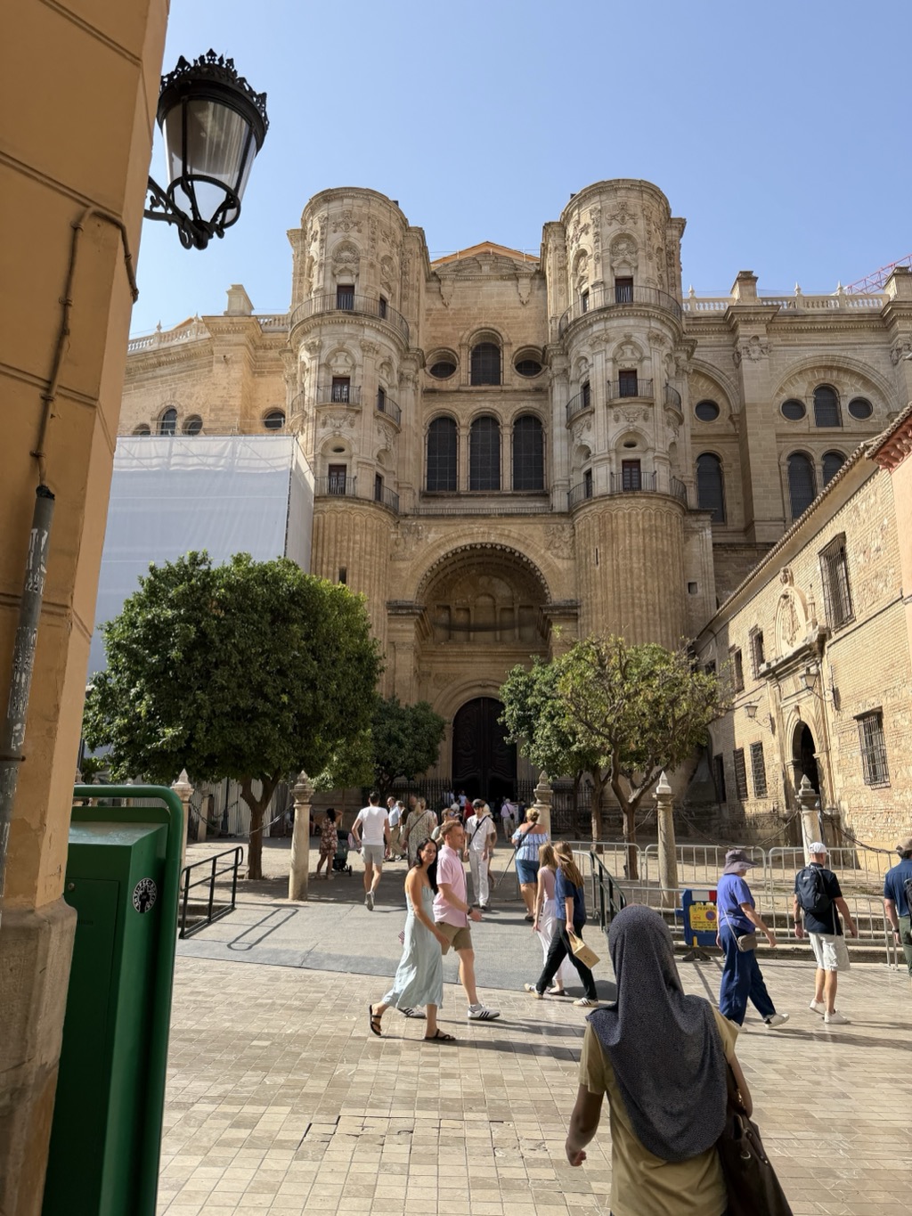Málaga Cathedral seen from a shaded side street, its honey-coloured stone facade rising above the plaza in full sun. Two heavily ornamented bell towers flank a tall central section of round-arched windows and carved pilasters, with a deep arched doorway below and white scaffolding sheeting across part of the left side. Orange trees stand in the square in front, and a mixed crowd of visitors in summer clothes -- sun hats, sundresses, shorts, one woman in a grey headscarf in the foreground -- crosses the pale paving. An ornate black iron street lamp hangs from the ochre wall at the left.