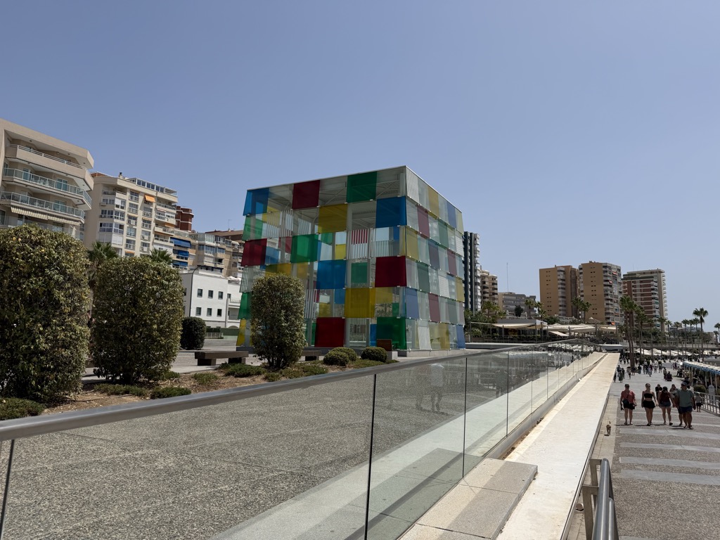 The Centre Pompidou Málaga on the harbour promenade -- a large cube of glass panels in red, yellow, blue, green and clear, sitting low against the skyline. A glass balustrade runs along the raised walkway in the foreground with clipped shrubs beside it, and the promenade below is lined with palms and white parasols where a scattering of people walk in the sun. Apartment blocks and hotels rise behind under a cloudless pale blue sky.