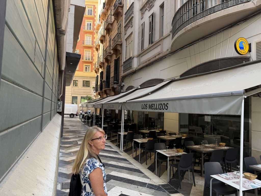 Sherri in a blue and white floral top and glasses walks along a narrow city street in shade, a black backpack over one shoulder. To the right, cream awnings lettered LOS MELLIZOS shade a row of pavement tables set with wine glasses and bread baskets, with a round yellow sign for the restaurant mounted on the wall above. The pavement is laid in black and white chevron marble, and tall ochre and cream apartment buildings with wrought-iron balconies rise on both sides under a narrow strip of bright sky.