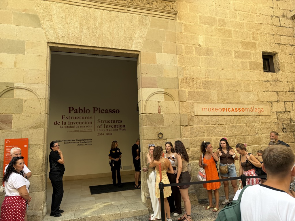 The entrance to the Museo Picasso Málaga, a deep stone doorway cut into a golden sandstone wall with museoPICASSOmálaga lettered on a plaque beside it in orange and grey. Inside the doorway a wall panel reads Pablo Picasso, Estructuras de la invención / Structures of Invention, Unity of a Life's Work, 2024-2028, with a second line for Joana Vasconcelos, Transfiguración. Two staff in black stand at the threshold and a queue of visitors waits behind a rope barrier at the right, mostly young women in summer dresses and shorts, some fanning themselves in the heat.