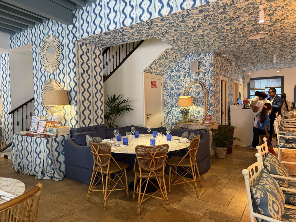 The dining room of Sushita Chinitas, decorated almost entirely in blue and white. Ikat-patterned fabric covers the walls and is gathered across the ceiling, with a large toile-papered wall behind a curved navy banquette. A round marble-topped table is laid with blue glassware and white plates, ringed by rattan cane chairs on a travertine tile floor. Starburst mirrors and a gilt-framed mirror hang on the walls, table lamps with linen shades stand on a skirted side table stacked with books, and potted palms fill the corners. At the right, staff and a family wait near the host stand by the front windows.