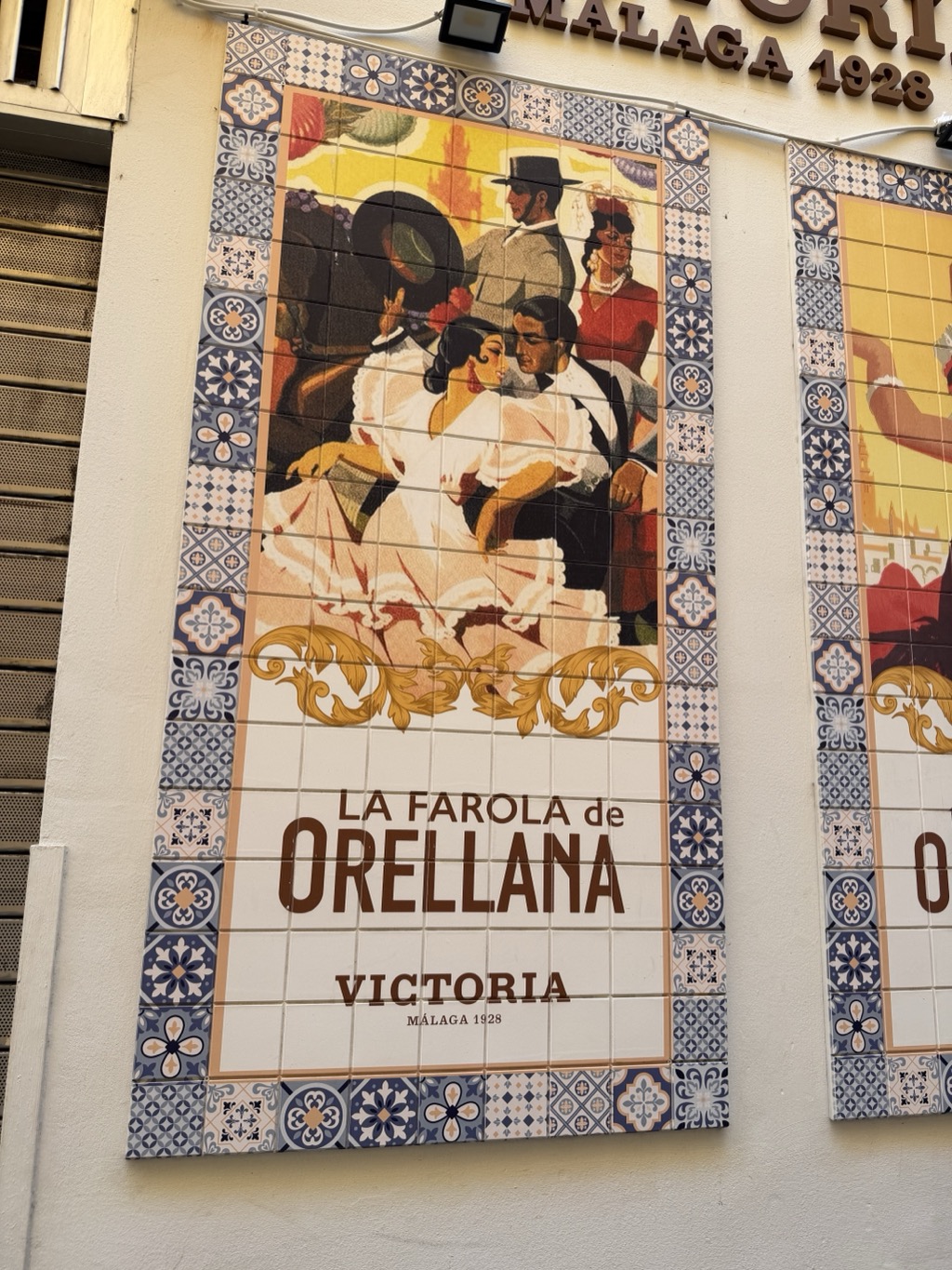 A large painted ceramic tile panel mounted on a pale wall, advertising Victoria beer. The artwork shows a flamenco scene in 1920s poster style -- a woman in a white ruffled dress dancing with a man in dark clothes, a figure in a wide black hat behind them, and a woman in red with a mantilla -- above gold scrollwork and the words LA FAROLA de ORELLANA, VICTORIA, MÁLAGA 1928. A patterned blue and white tile border frames the panel, a second panel is visible at the right, and raised lettering reading MÁLAGA 1928 runs along the wall above.