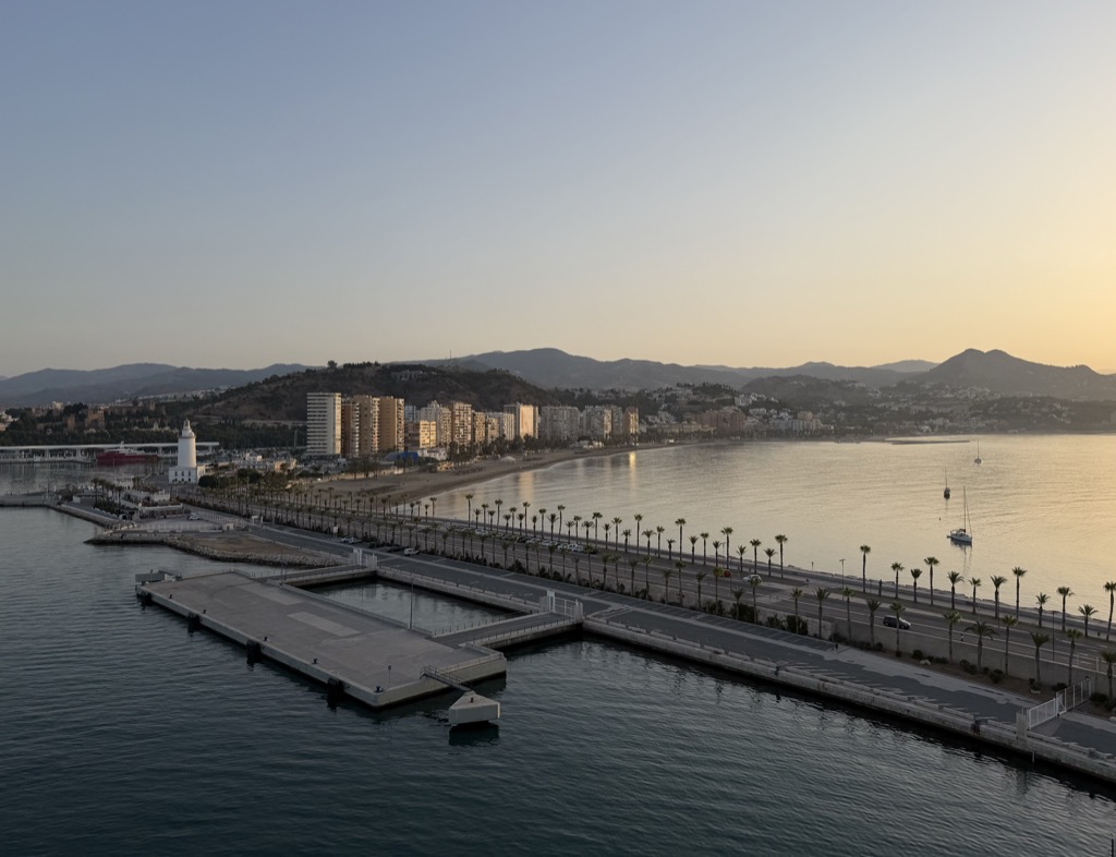 The Málaga waterfront seen from high on the ship in early morning light. A long concrete quay and a palm-lined promenade run across the frame toward the white La Farola lighthouse, with the curve of La Malagueta beach behind and rows of apartment blocks along the seafront. Dry brown hills rise beyond the city under a pale sky washed gold near the horizon, and a couple of sailboats lie at anchor on flat water at the right.