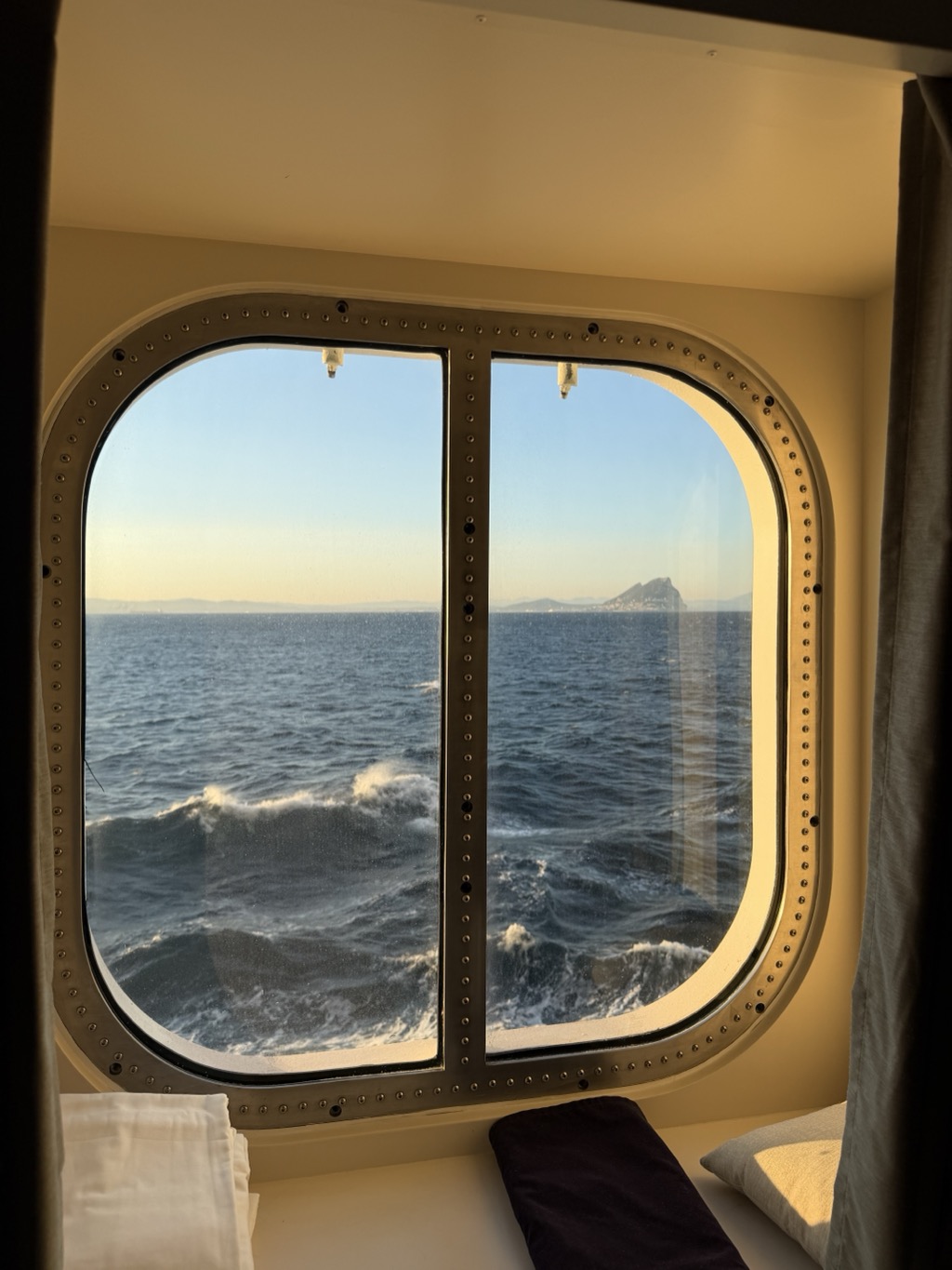 The Rock of Gibraltar seen through a cabin window at golden hour, its distinctive ridge silhouetted low on the horizon across open water. The window is a large rounded rectangle set in a cream frame studded with rivets and divided by a central bar, with a grey curtain drawn back at the right. Below the glass, white bedding and a dark blue runner are visible on the window seat. The sea is a deep rippled blue with whitecaps breaking in the ship's wake, and the sky above fades from pale blue to warm gold.
