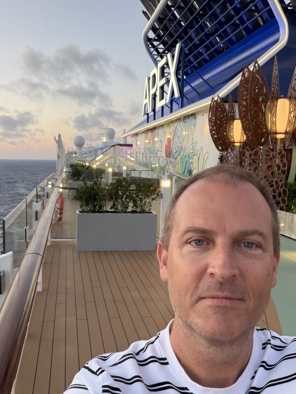 A selfie of Brian on the Celebrity Apex upper deck at dusk, his face filling the right of the frame. He has short light brown hair and blue eyes and wears a white t-shirt with irregular black horizontal stripes. Behind him the teak deck runs forward along the port rail past a white planter of clipped green shrubs, with warm deck lights already lit. The ship's blue funnel rises at upper right lettered APEX in white, beside a wall of tall perforated rust-coloured metal screens and a mural in pale blues and yellows. The flat Atlantic is dark at the left under a pale sky streaked with pink and grey cloud near the horizon.