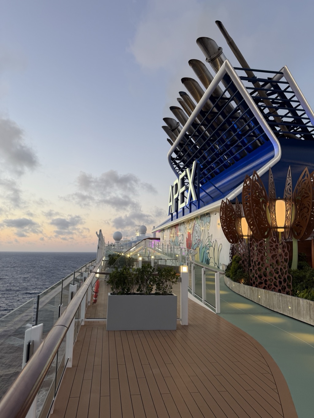 The upper deck of the Celebrity Apex at dusk, looking forward along the port rail. The ship's blue funnel housing rises at the right with APEX lit in white letters across it and a bank of tall exhaust stacks angled behind a black lattice screen. Below it, a curved wall is painted with a colourful mural, and a row of tall rust-coloured cut-metal lanterns glows on a raised planter bed. Teak decking and a glass balustrade run away toward the bow, past a planter of clipped greenery and a red life ring, with the dark Atlantic to the left and a pale pink and blue sunset sky along the horizon.