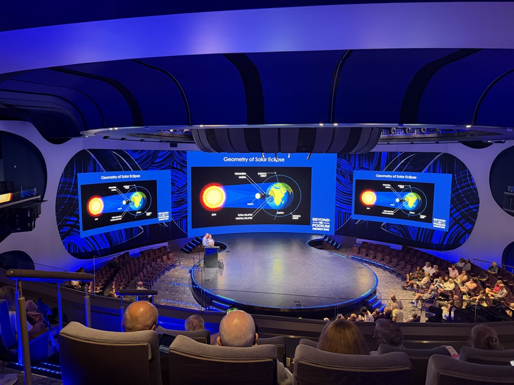 The ship's theatre during a daytime lecture, shot from the back of the raked seating. Three curved screens across the stage wall all show the same slide titled Geometry of Solar Eclipse, with a diagram of the sun, moon and Earth on a black background. The room is lit deep blue, with sculpted white ceiling ribs and a balcony rail curving overhead. A speaker in light clothing stands at a lectern beside the round stage, which is edged with blue LED strip. Perhaps a hundred passengers sit watching in grey armchairs, more filling the seats along the right-hand side.