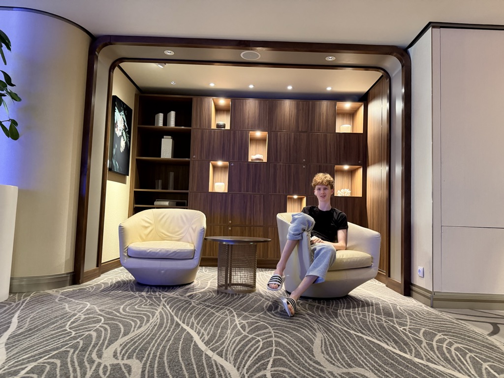 Rowan sits sideways in a cream leather tub chair in a quiet corner of a ship's lounge, legs draped over one arm of the chair, wearing a black t-shirt, pale blue sweatpants and black slides, looking toward the camera. A dark walnut display wall of open shelves fills the space behind him, lit from within and holding vases, books and ornaments. A second matching chair and a small round mesh side table sit at the left, a framed dark floral picture hangs on the wall, and the floor is laid with a grey carpet in a swirling contour pattern.