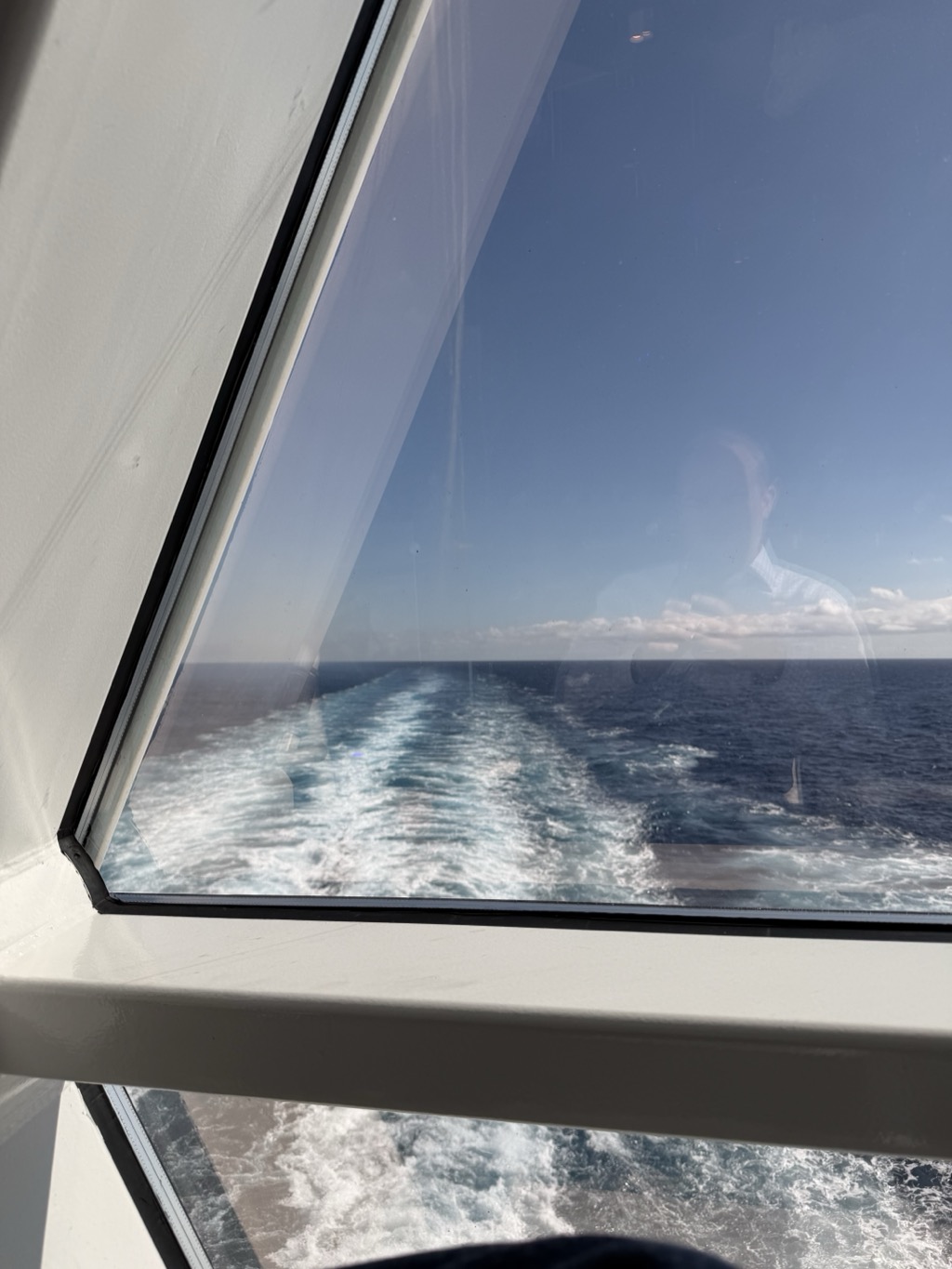 The ship's wake seen through a large angled window, the white-painted frame cutting diagonally across the top left of the frame. Beyond the glass, a broad churning band of white water trails away to the horizon across deep blue open ocean, under a clear blue sky with a line of small white clouds low on the horizon. A faint reflection of a person is visible in the glass at the right, and a second pane below the sill shows more of the wake.