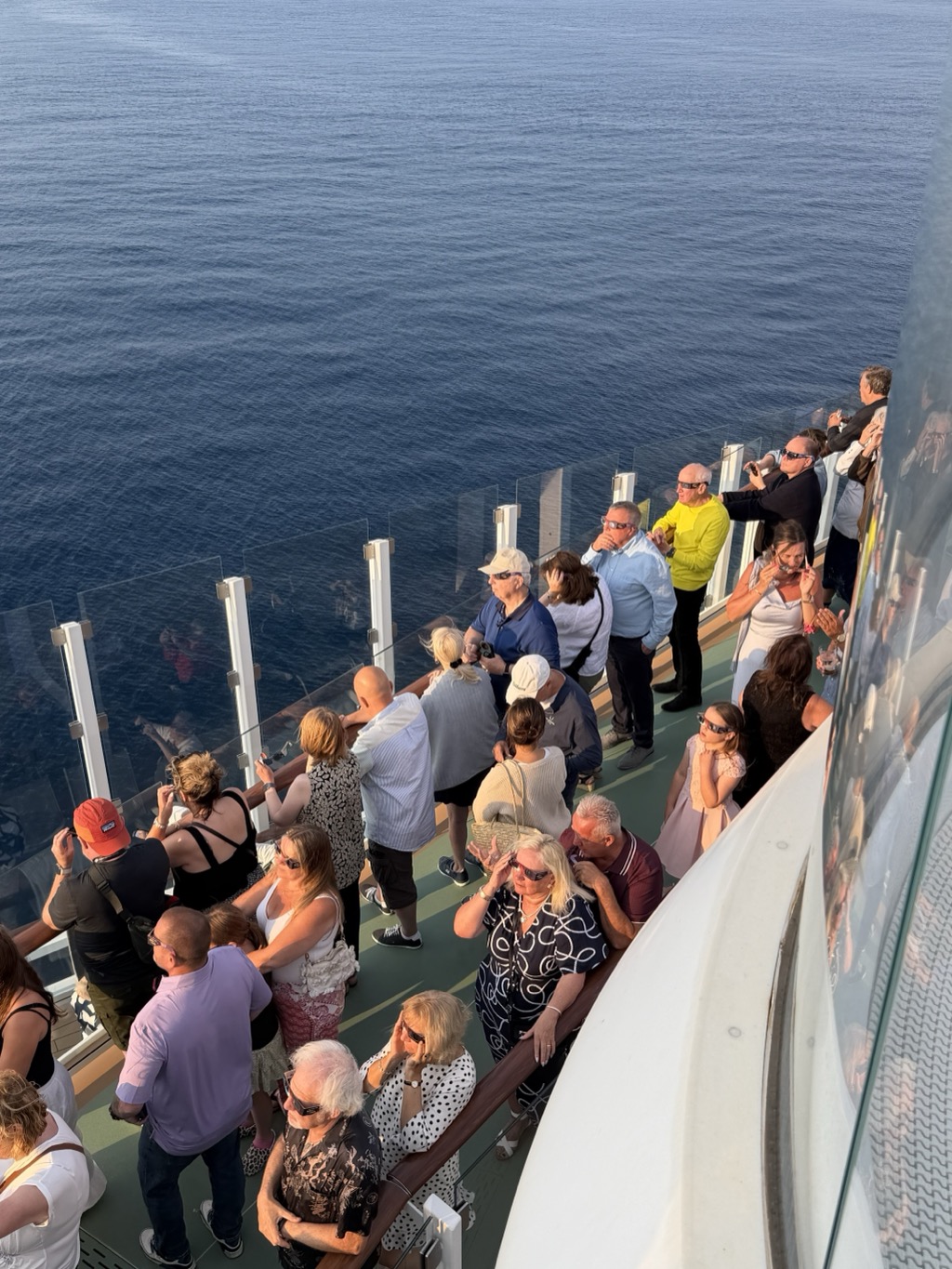 Looking steeply down from an upper deck onto a crowded outer deck of the ship in warm low evening light, with the flat dark blue Atlantic filling the left half of the frame beyond a glass balustrade. Dozens of passengers are packed along the green-striped deck and the timber handrail, nearly all wearing dark cardboard eclipse glasses and tilting their faces up toward the sun out of shot. Among them are a man in a bright yellow jumper, a man in a pale blue shirt, a woman in a white dress, a young girl in a pink dress and several people in caps and sun hats; some hold phones up to photograph the sky. A curved white section of the ship's superstructure and a large tinted window cut across the right of the frame, reflecting more of the crowd.