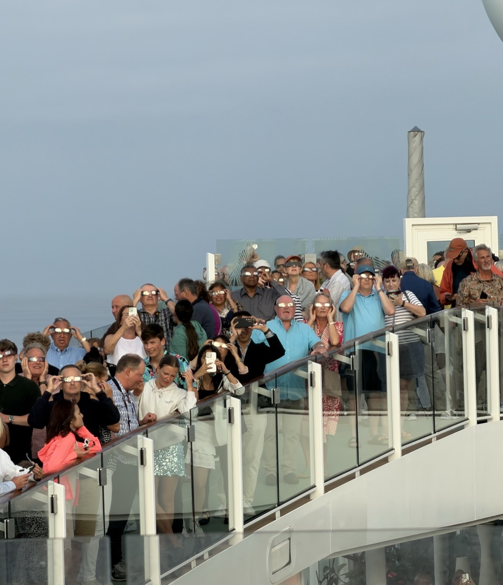 Passengers packed along the curved glass balustrades of the ship's upper decks during the partial phase, nearly all of them wearing cardboard eclipse glasses and tilting their faces up toward the sun out of shot. The lenses catch the light as bright rectangles across dozens of faces. Several people hold phones up to photograph the sky instead. The crowd is in summer clothes — a man in a turquoise polo, a woman in a coral jacket, a girl in a white sweatshirt and sequinned skirt, a man in an orange cap — and stands two tiers deep along the curve of the rail. A white ventilation mast and a corner of the superstructure rise at the right, and the flat pale sea meets a hazy twilight sky behind them.