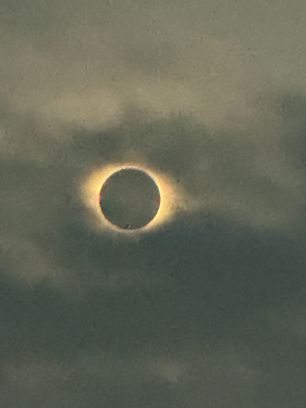 Totality, photographed through a veil of thin grey cloud. The moon sits as a flat dark disc squarely in the centre of the frame, ringed by a narrow band of pale golden light from the sun's corona, brightest along the left and right edges where faint flecks of red and orange show. The glow diffuses outward into a soft halo before fading into the surrounding sky, which is an even dull olive grey mottled with darker drifting cloud. No landscape or horizon is visible.