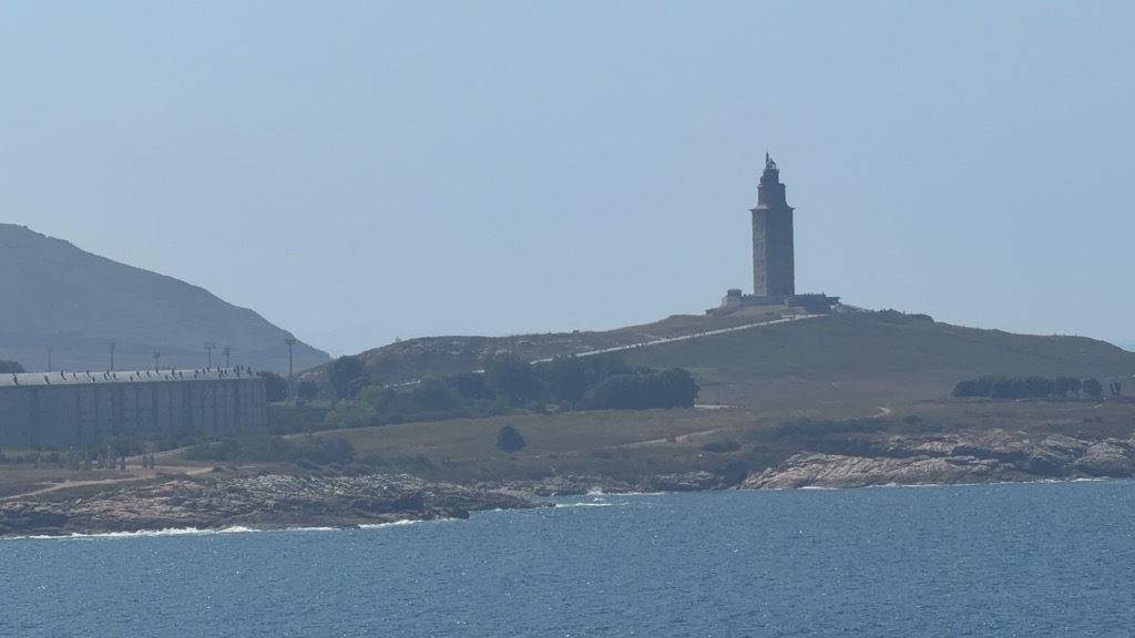The Tower of Hercules seen from the water on departure, across a stretch of deep blue sea with small whitecaps breaking on a low pink granite shoreline. The square Roman lighthouse tower stands in silhouette on the crown of a bare grassy headland, its stone warm brown in the haze, topped by a smaller lantern stage and cupola. A pale path traces the ridge of the headland below it and tiny figures are dotted along the skyline. To the left, a long low modern building with a heavily ribbed concrete facade runs along the shore beneath a hazy hill, with a line of pylons and scattered trees between. The sky is a pale washed-out blue.
