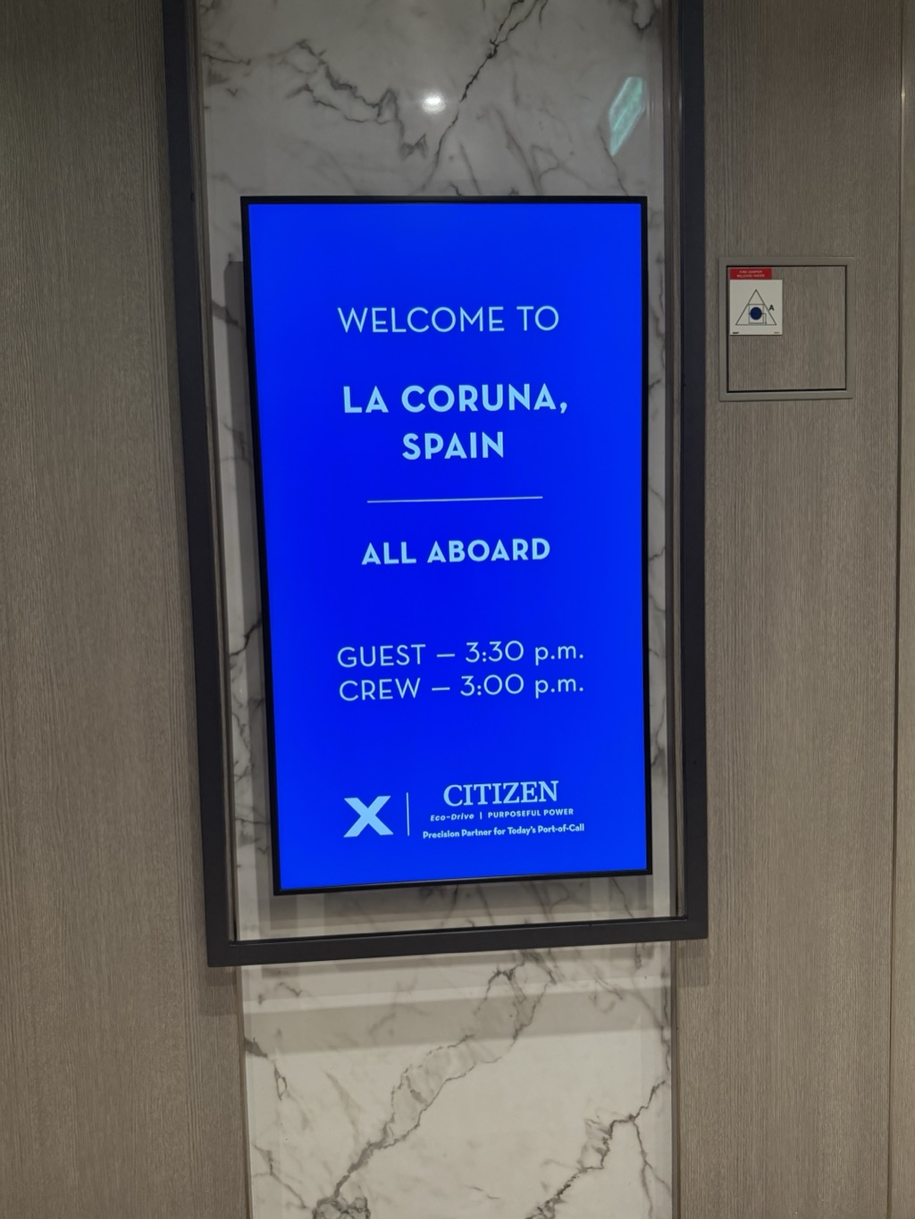 A vertical digital signboard set into a marble-and-timber wall in a ship's elevator lobby, glowing bright blue. White text reads WELCOME TO LA CORUNA, SPAIN, then below a dividing rule, ALL ABOARD, then GUEST — 3:30 p.m. and CREW — 3:00 p.m. At the bottom is the Celebrity X logo beside CITIZEN, Eco-Drive, PURPOSEFUL POWER, and the line Precision Partner for Today's Port-of-Call. A small red fire-damper inspection plate is mounted on the pale wood panelling to the right.