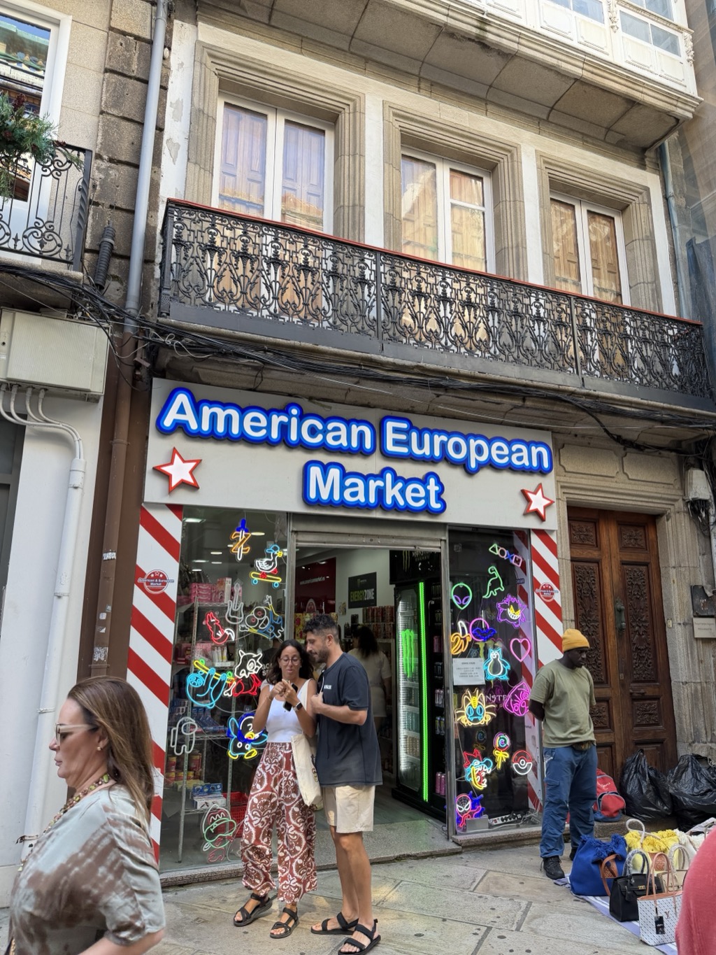 A shop called American European Market on a narrow old-town street, its name in blue outlined lettering on a white fascia flanked by two red and white stars, with red and white candy-striped pilasters either side of the open doorway. The windows are covered in brightly coloured neon-style outline decals of cartoon characters, game controllers, an alien head and a heart. Inside, shelves of colourful packaged sweets and drinks are visible along with a glass-fronted cooler lit bright green. Above the shop is a grey stone facade with a long black cast-iron balcony in an elaborate scrolled pattern and tall shuttered windows. Several people stand on the pavement: a couple in the doorway, the woman in a white vest and patterned wide-leg trousers looking at something in her hands, the man in a dark t-shirt and beige shorts; a man in an olive t-shirt and mustard beanie by a heavy carved wooden door at the right, beside black bags and a display of handbags laid out on the ground; and a woman in a brown tie-dye top in the near foreground.